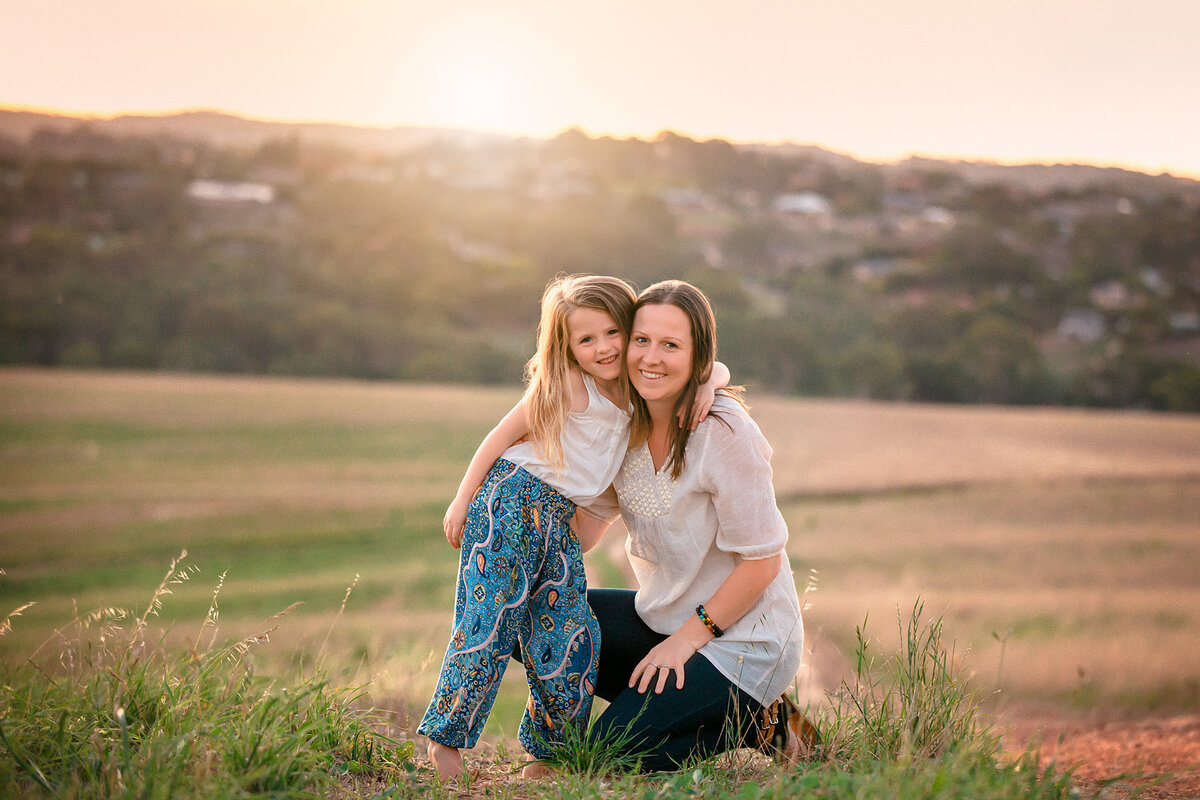 Family photographer Hampton Roads va photographs mommy and me session with child hugging her mother snack as the mother crouches down next to her and the sunshine's over a hill in the distance