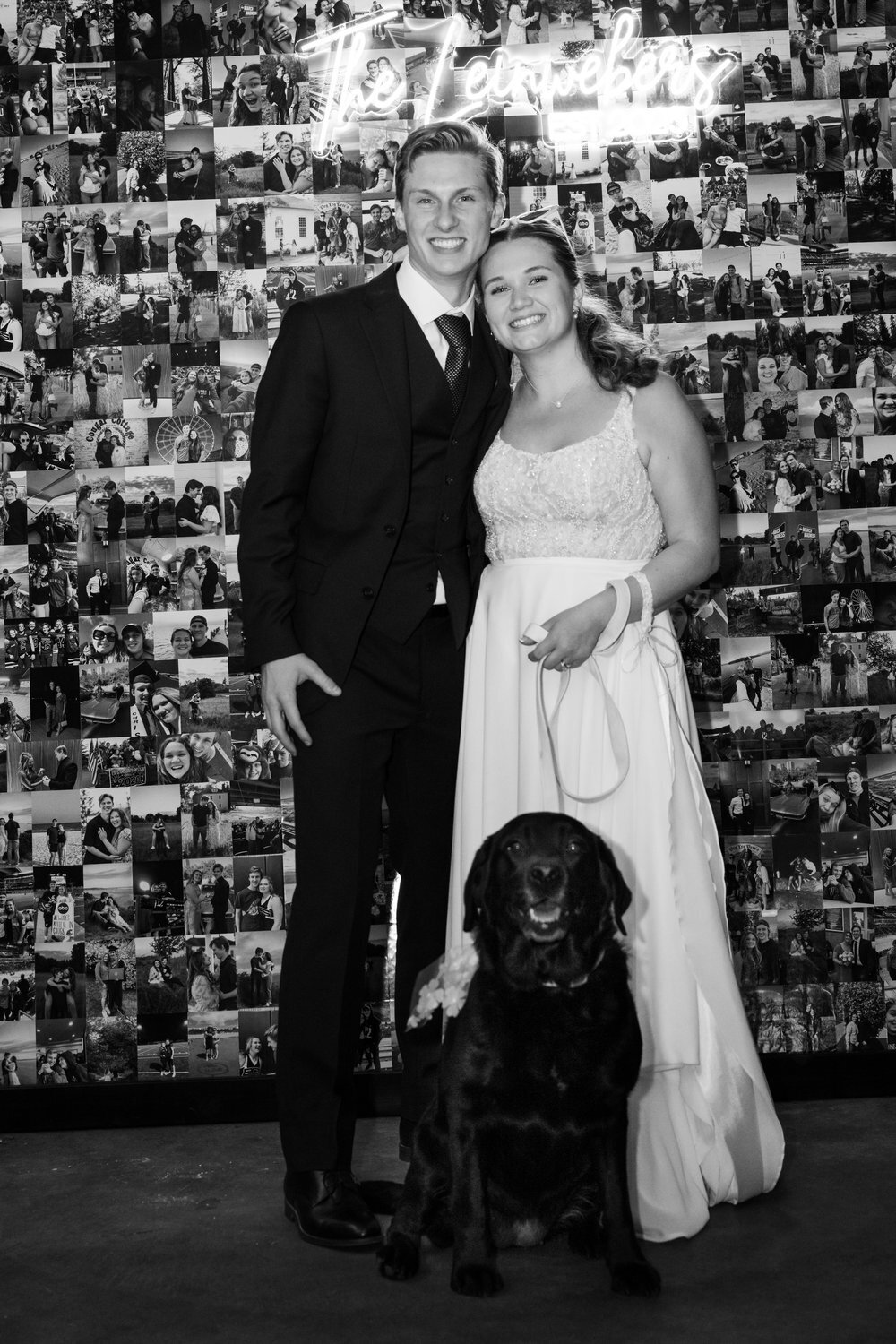 black and white image bride and groom with black lab in front of photo wall at reception