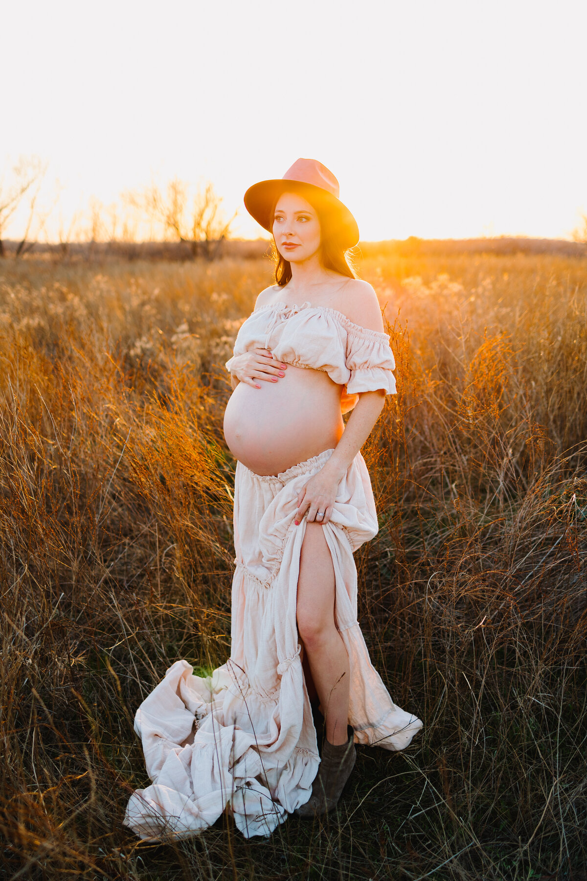 Photograph of pregnant woman in a garden with tall grass, she is showing her belly dressed in a skirt and a short white t-shirt. On her head is a brown hat and she is looking to the left side.