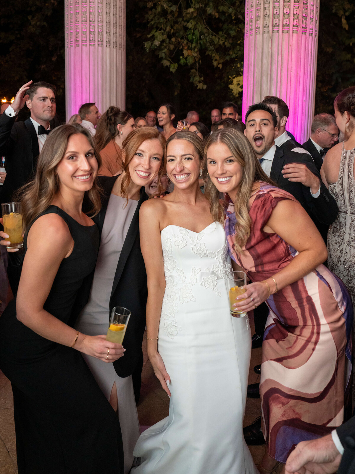 A smiling bride in a white dress poses with four women holding drinks at an event. They stand under columns illuminated by purple lights, with a crowd of people in formal attire in the background.
