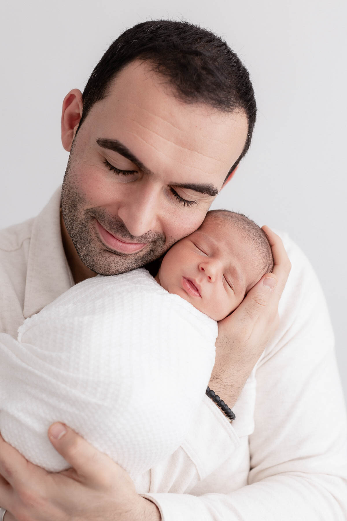 Dad holding newborn baby cheek to cheek. Dad has eyes closed and baby is sleeping. Dad is wearing a cream colored shirt and baby is wrapped in a white textured wrap.