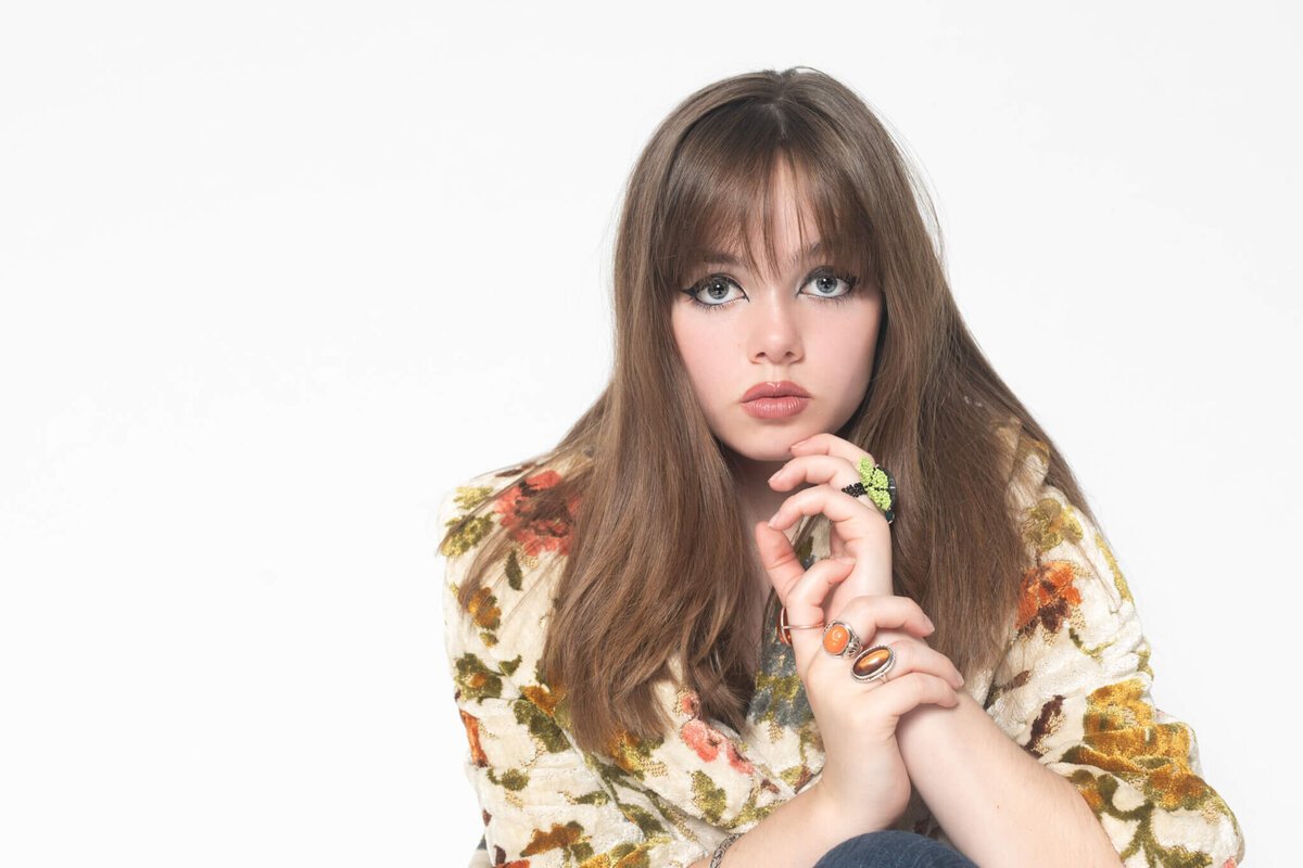 A beautiful model with long, brown hair and bangs sitting in a studio with her hand near her face, wearing a floral coat in a white studio in Tustin.