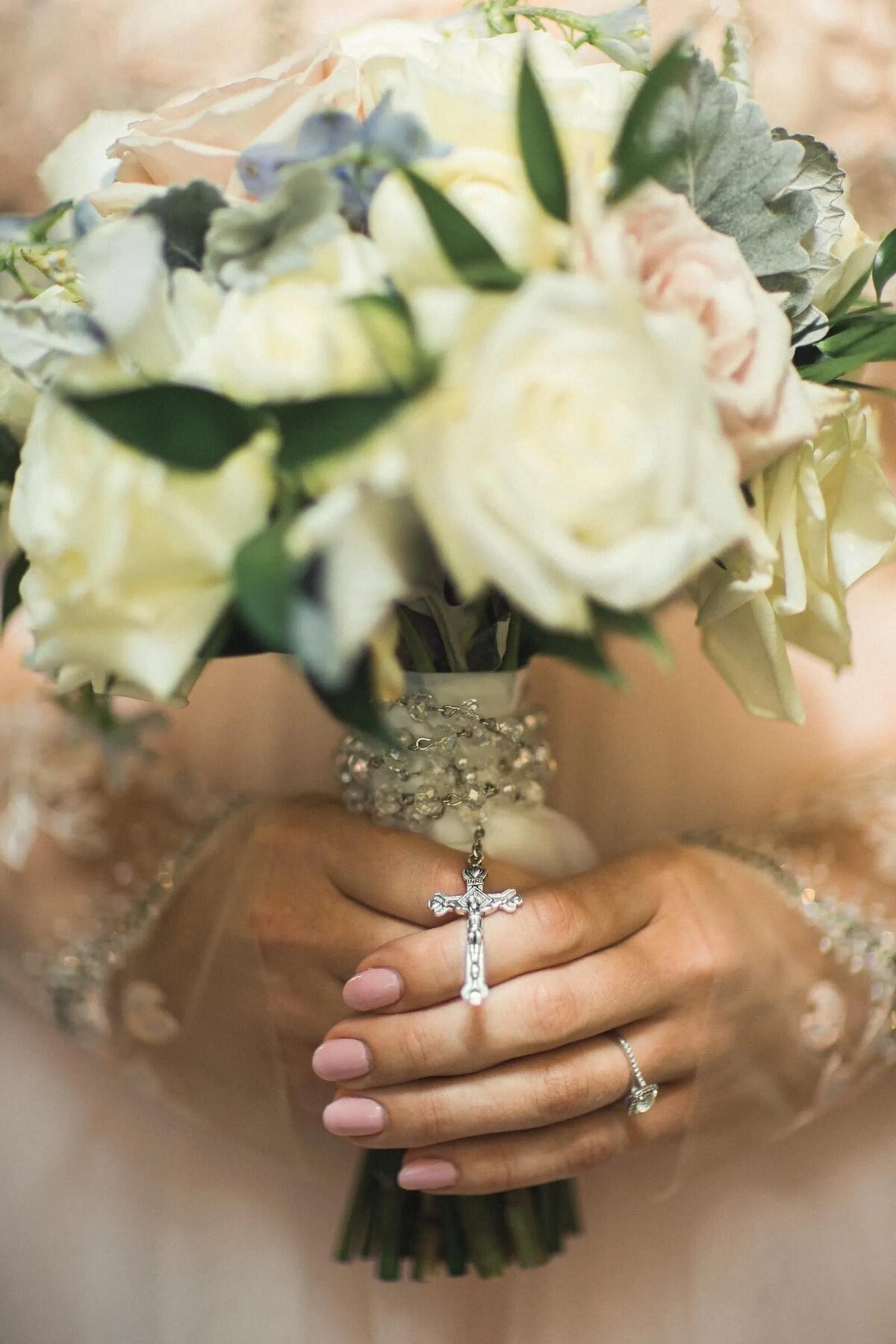A close-up of a bride's hands clasped over a bouquet, showcasing her wedding ring and a delicate cross