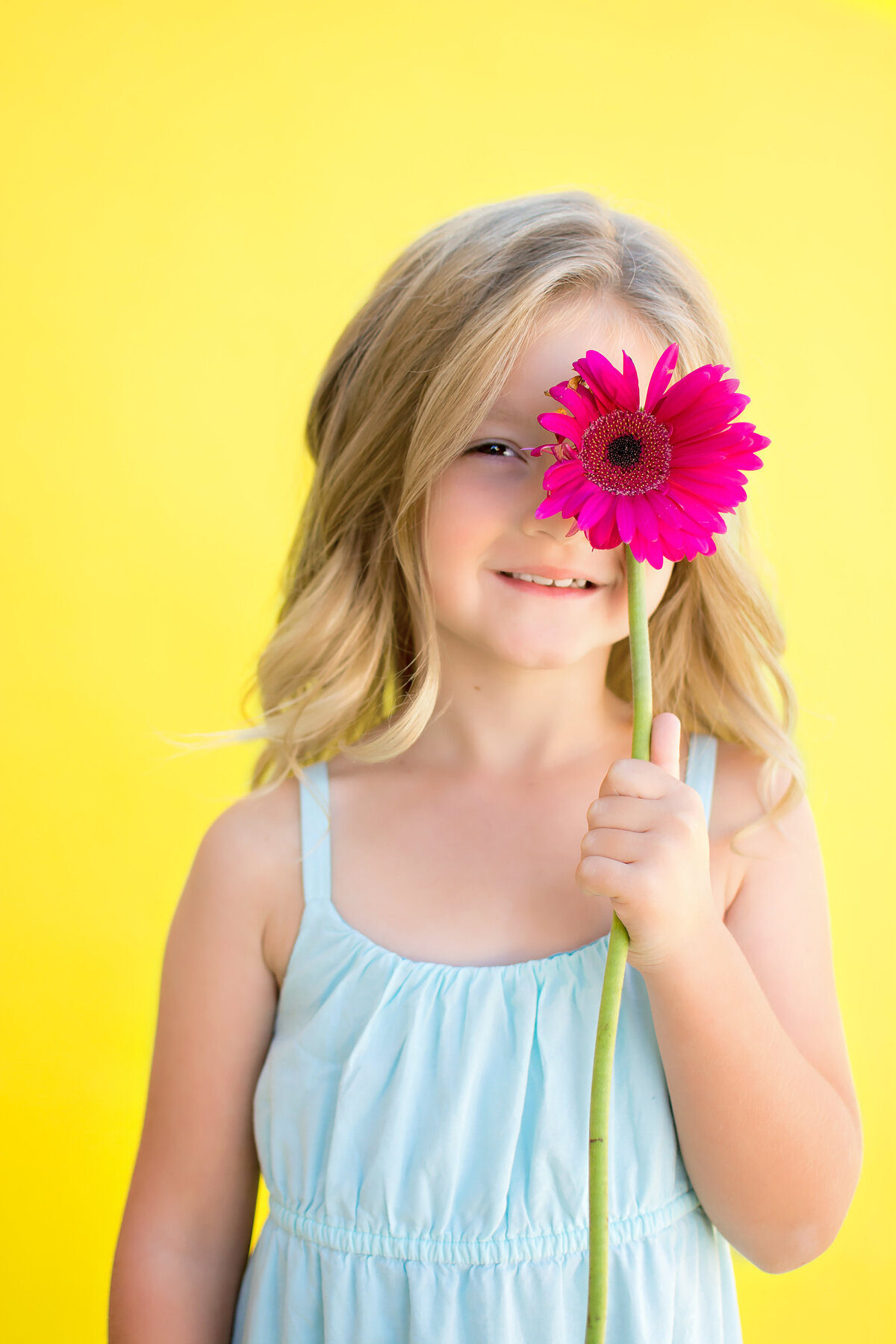 Young girl standing on yellow backgroun with blue dress and a bright pink flower in front of her eye.