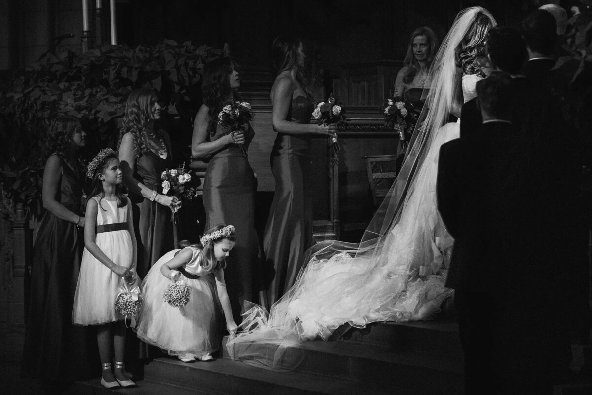 A black and white image of a young flower girl adjusting the bride's dress, with bridesmaids watching in the background