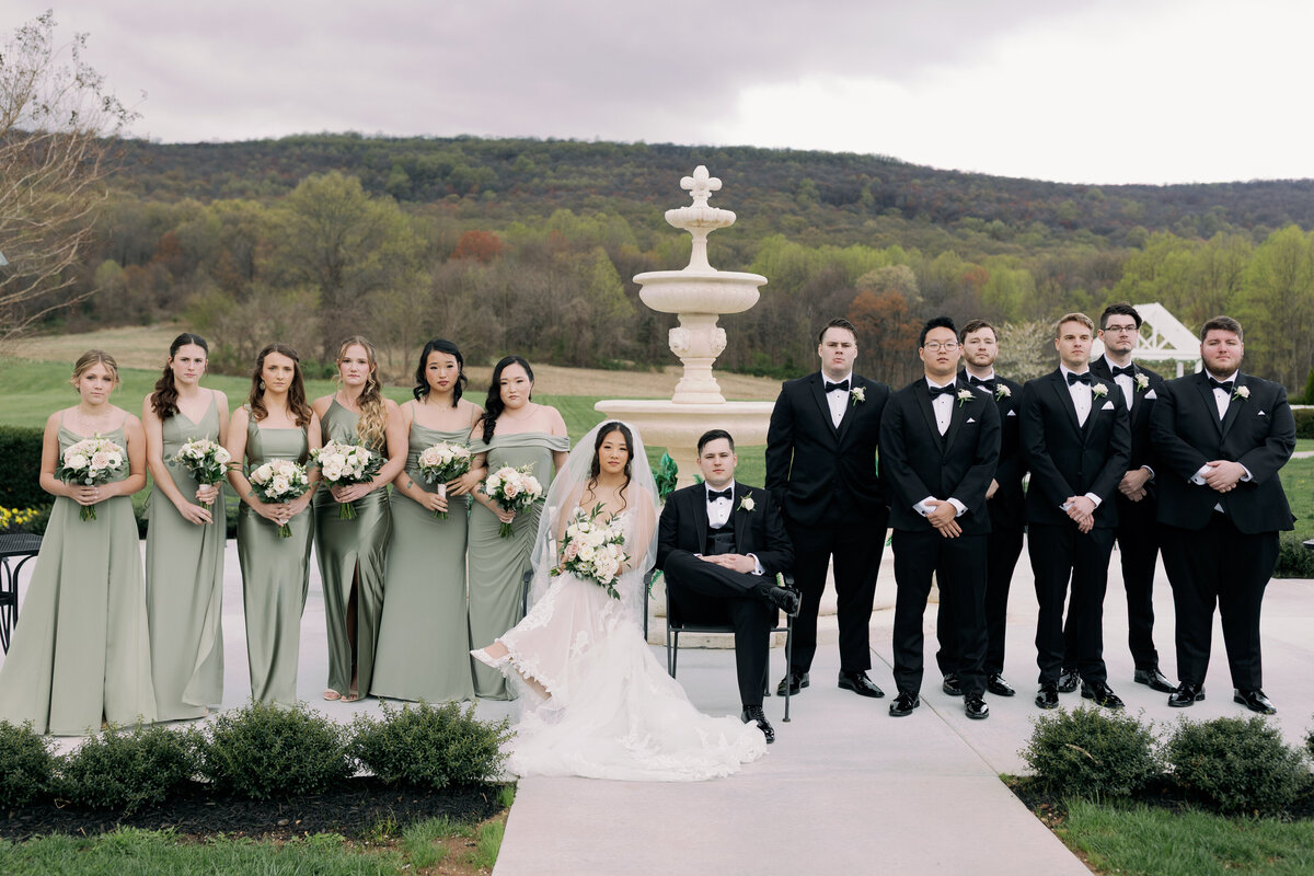 Bridesmaids in sage green satin dresses and groomsmen in black tuxedos posing by the Springfield Manor fountain.