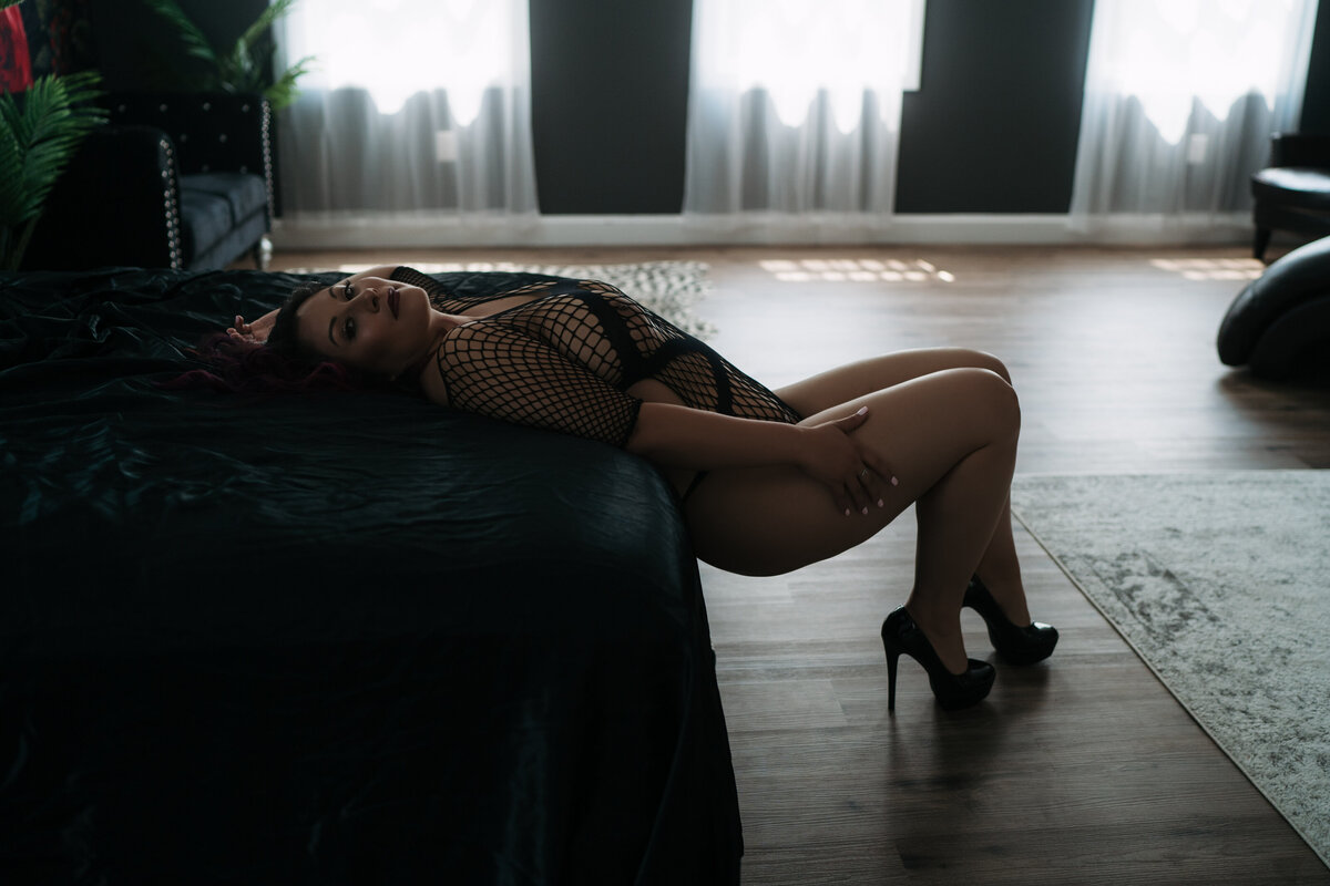 A woman in black mesh lingerie leans back on a black bed with her feet on the wood floor in front of windows in a studio