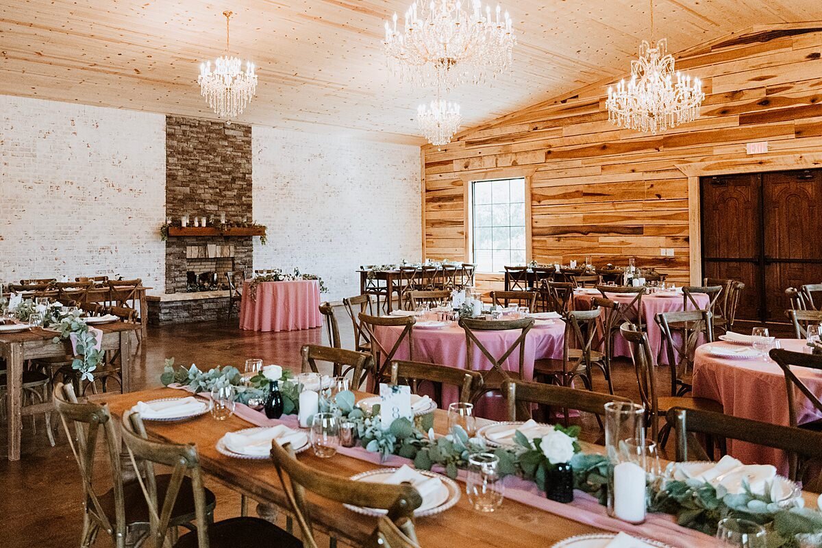 Long barn wood farm tables set with sheer blush table runners and long garlands of greenery and candles as well as round tables with blush table cloths fill the room. The tables are set with cross back barn wood chairs, gold chargers and white plates. Glass cylinders with pillar candles and white flowers decorate the tables. Crystal chandeliers sparkle from the ceiling inside Steel Magnolia Barn.