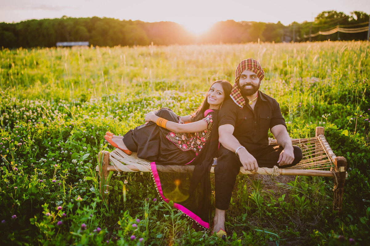 A sikh couple sitting on a manja in an open field in connecticut during their punjabi themed engagement session wearing traditional punjabi outfits
