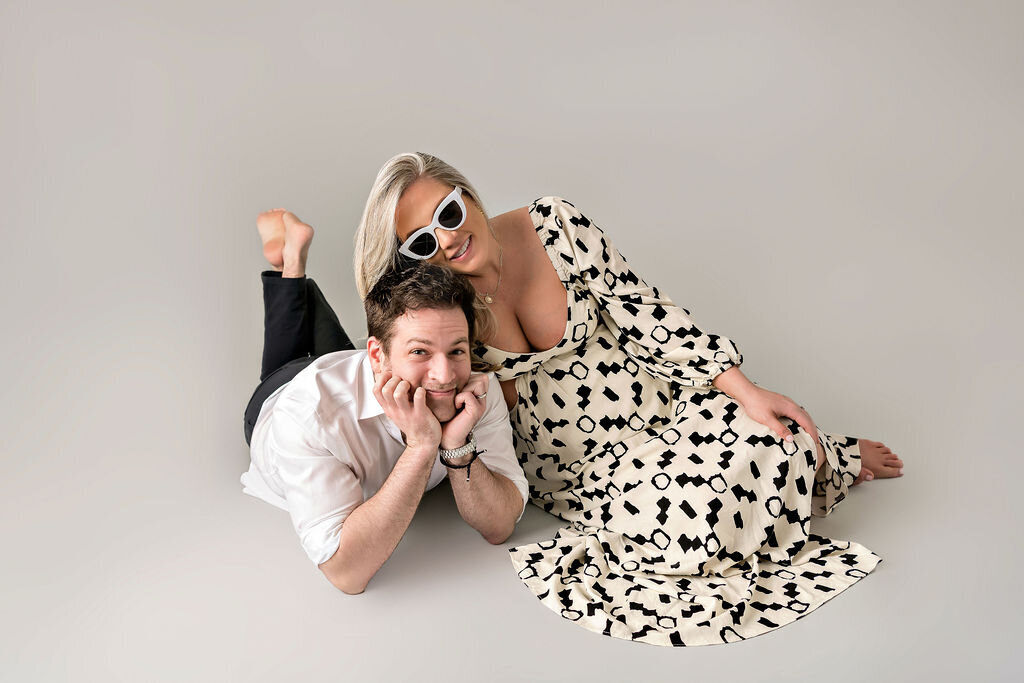 Playful studio maternity session with couple lying on floor in coordinated outfits