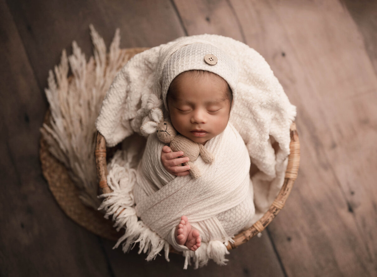 Newborn baby photoshoot; aerial image. Baby  boy swaddled in white waffle stretch fabric and matching hat is holding a tiny stuffed bunny. He is placed in a basket and his toes are peeking out of the wrap. Baby boy sleeping peacefully. Captured by best Lake Elsinore newborn photographer Bonny Lynn Photography.