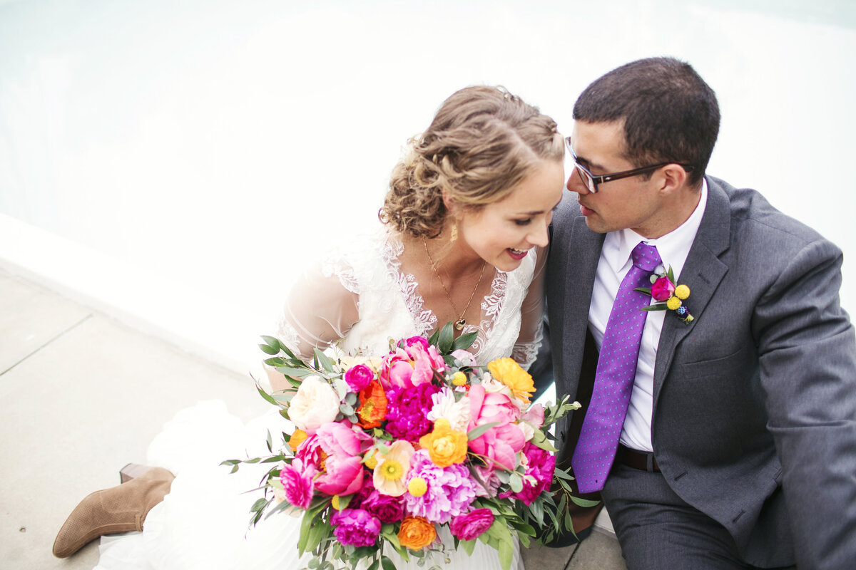 A joyful bride and groom share a sweet moment, with the bride holding a bold and colorful bouquet of pink, orange, and yellow florals.