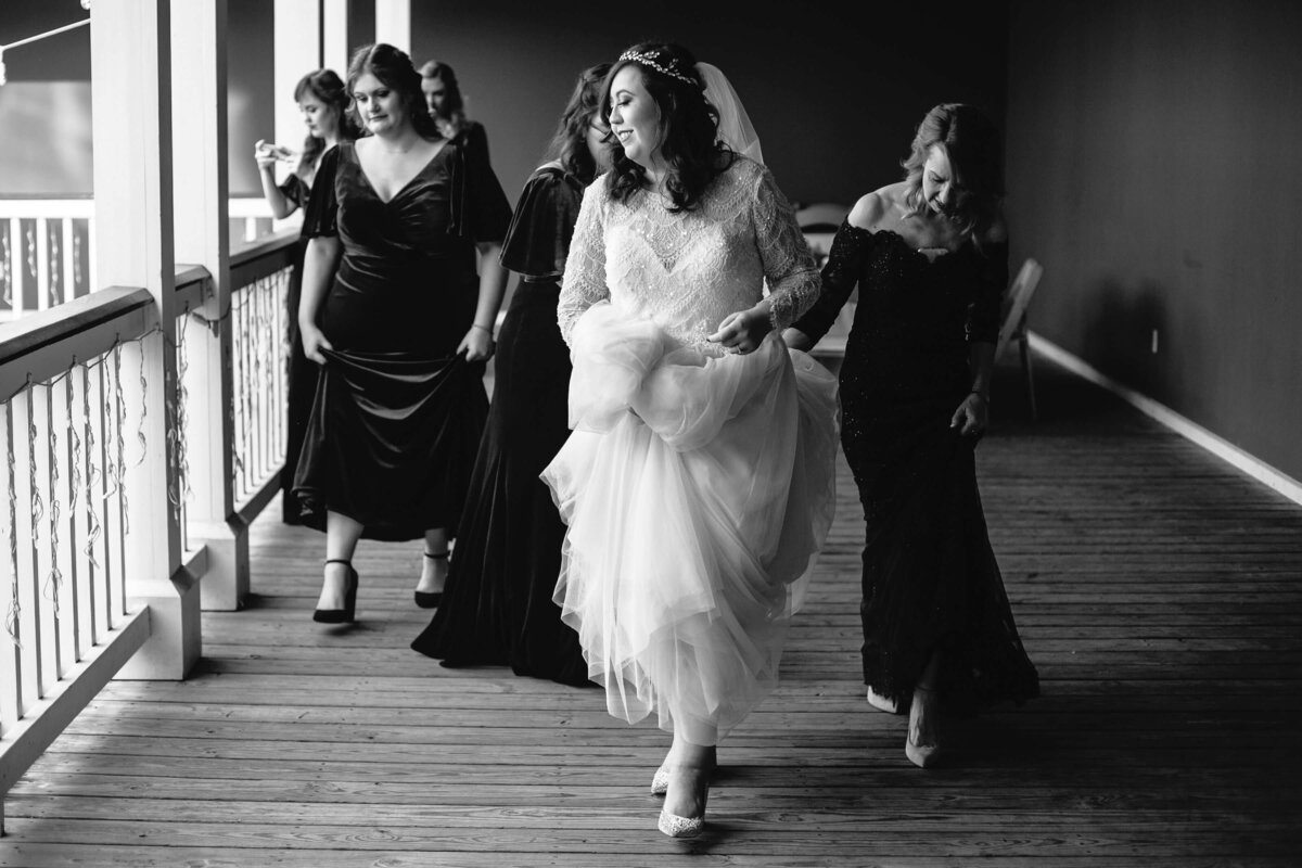 A bride in a white dress walks happily down an outdoor hallway, holding her gown, followed by bridesmaids in dark dresses. The scene is joyful and candid, with natural light streaming in from the left.