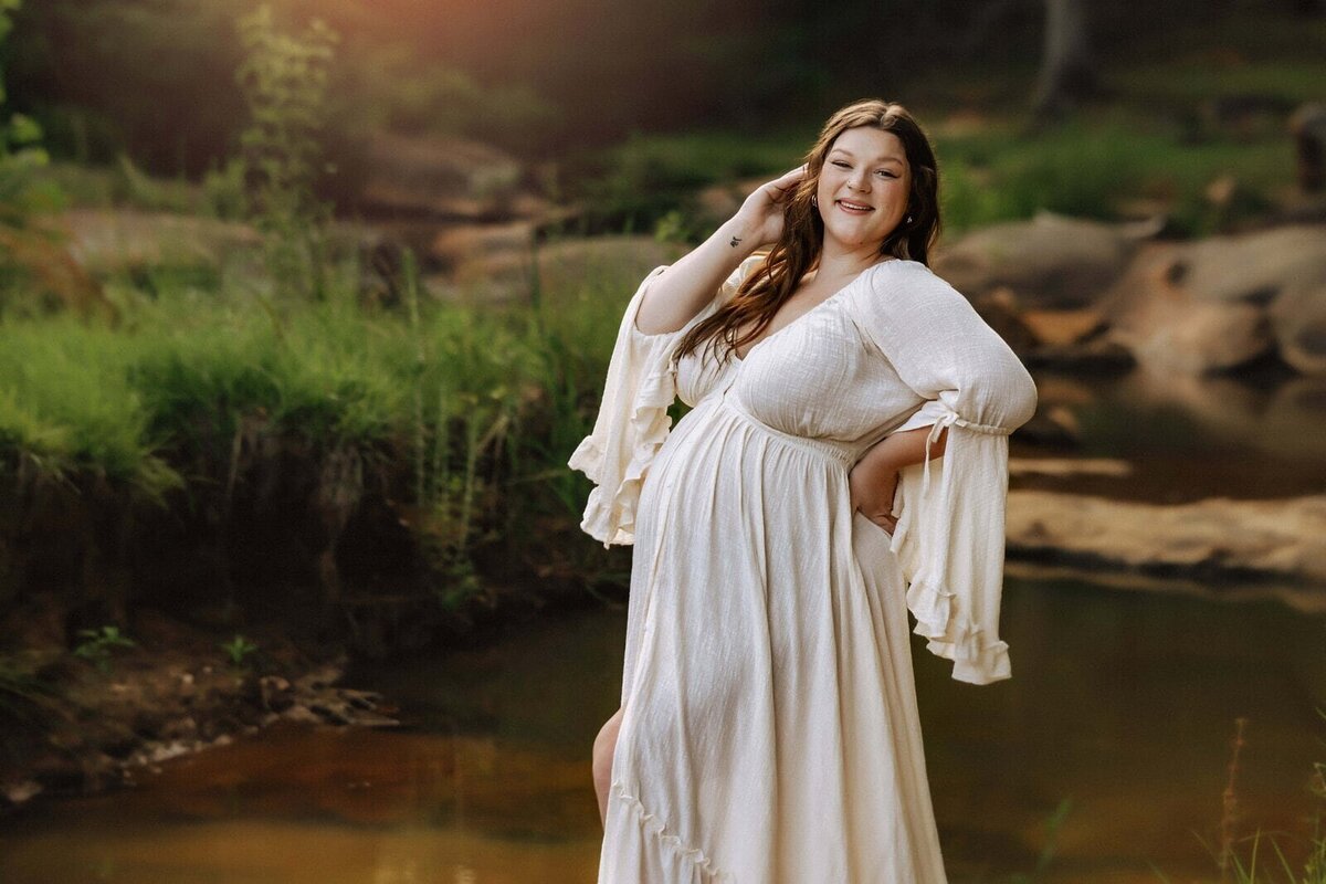 A person in a white dress poses near a calm stream in a forested area, with greenery in the background and soft sunlight filtering through.