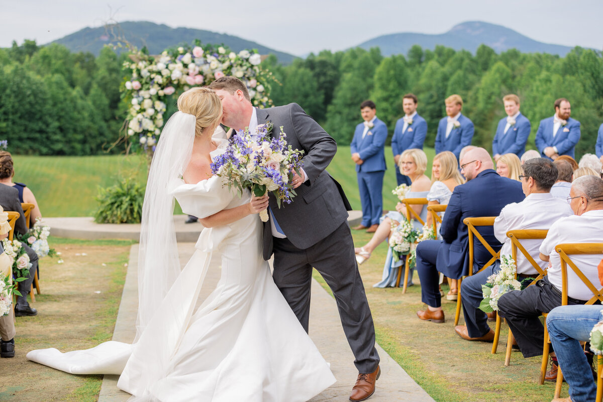 Bride and groom share a kiss as they walk down the aisle with the beautiful mountains setting the perfect backdrop for a summer wedding. 