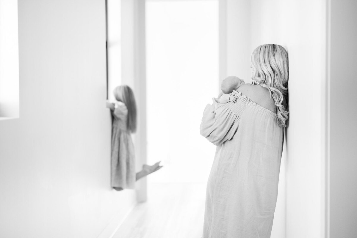mom holding baby watching big sister look out the window for Tampa newborn photographer Tina Marie Studio.