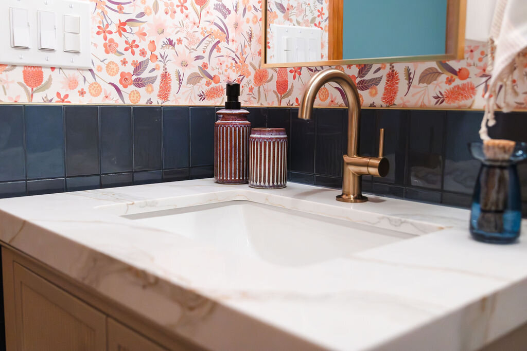 A close-up of a bathroom vanity featuring a white Calacatta Gold Quartz countertop with subtle veining. A rectangular undermount sink is fitted with a modern brass gooseneck faucet. Several decorative items sit on the countertop: a tall, ribbed brown glass bottle with a black pump, a shorter, ribbed metallic brown tumbler, and a small blue glass bottle with a cork stopper. The backsplash is made of dark blue vertical tiles. The wall above the tiles is covered with a vibrant floral patterned wallpaper in shades of pink, orange, and white. A partially visible wooden-framed mirror hangs above the sink. Light switches are visible on the wall to the left. A glimpse of a white towel with tassels is on the right.