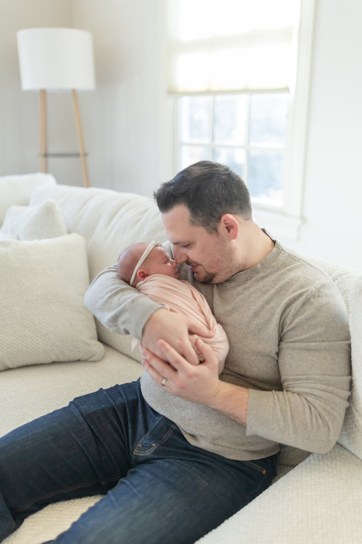 Dad gently holding his newborn baby while sitting on a comfortable couch in their home during an in-home session by Connecticut Newborn Photographer Oak and Lore Studio. The soft lighting and cozy atmosphere capture a tender, intimate moment between father and child