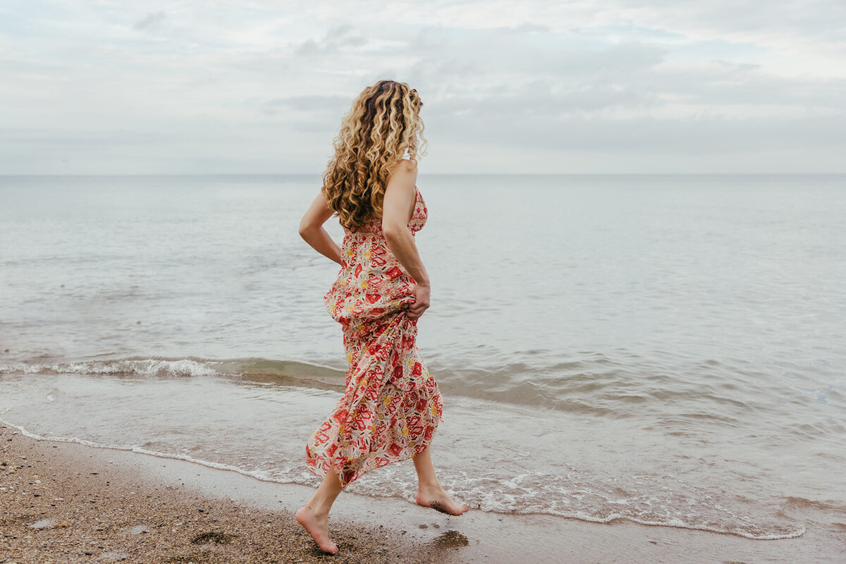 woman in long red floral dress is barefoot and stepping on her tiptoes into Lake Michigan