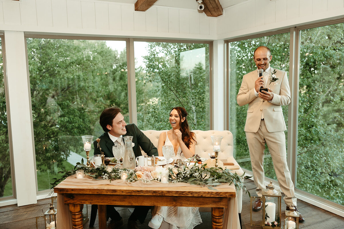 bride and groom sitting at a sweetheart table at Willowbrook wedding venue while a  father gives a toast image