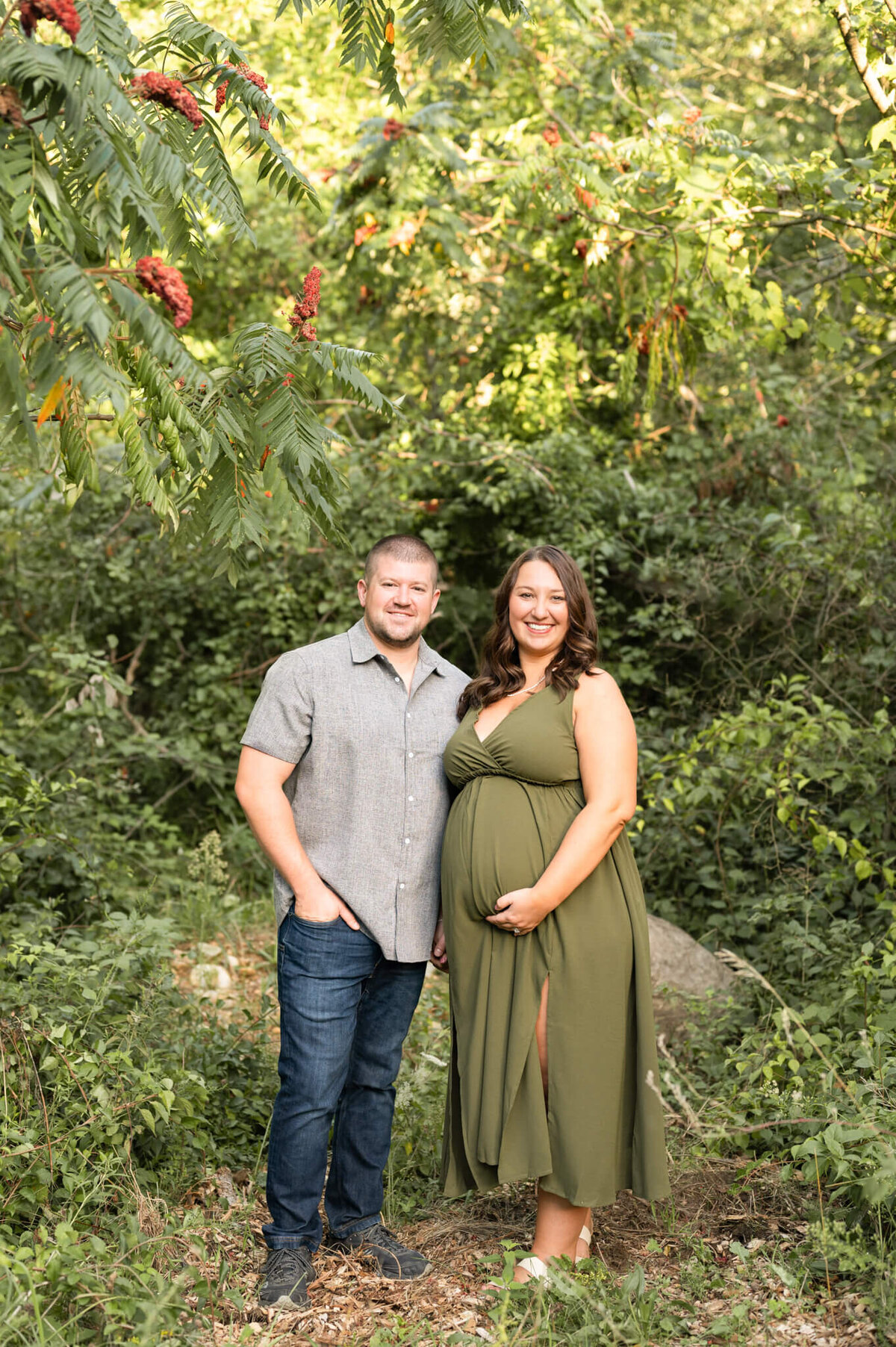 husband and wife posing for maternity photos in a forest surrounded by sumac