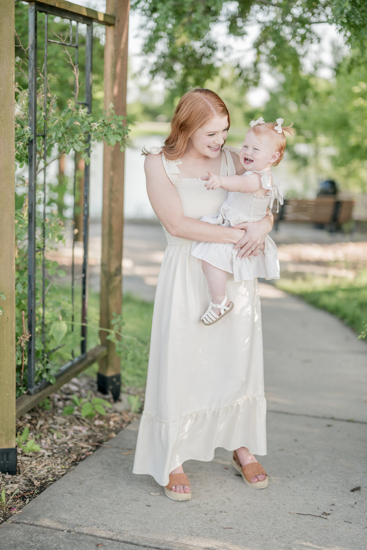 Mother holding her toddler in a light-filled outdoor setting with a fenced garden path in the background.