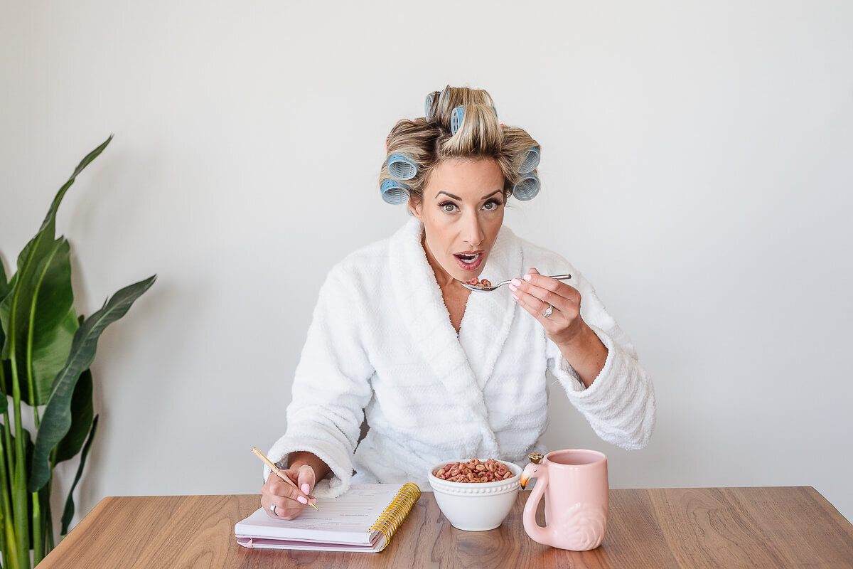 Blonde woman in hair curlers and bathrobe eating a bowl of cereal writing in notebook with coffee nearby
