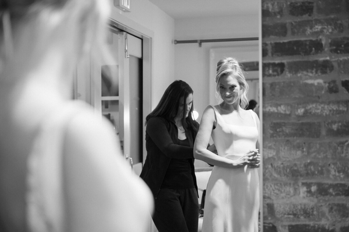 bride getting ready in bridal suite at common house richmond weddingvenue with her bridesmaid helping her get into her dress captured by Richmond wedding photographer