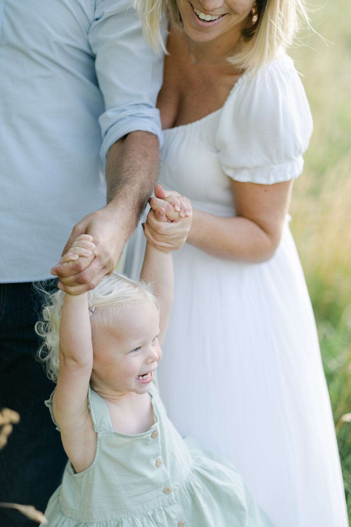 Artistic family photography of a mother and father holding their daughter's hands in a field at sunset