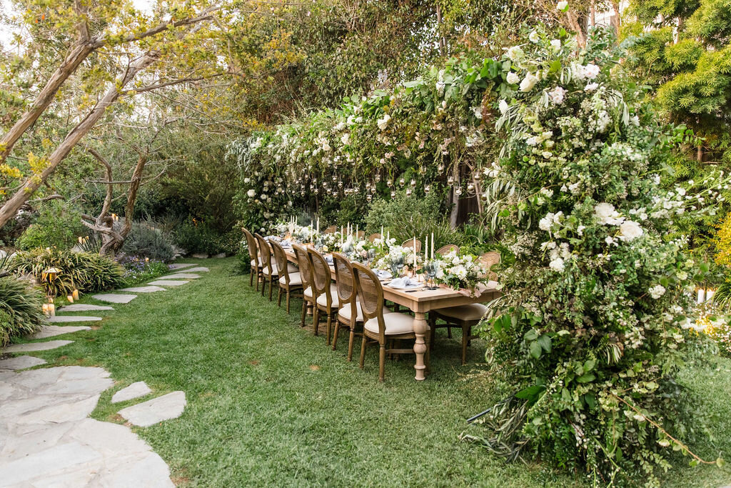 Intimate outdoor wedding reception set up featuring one long grand wooden table, with a huge floral arch spanning the length of the table.