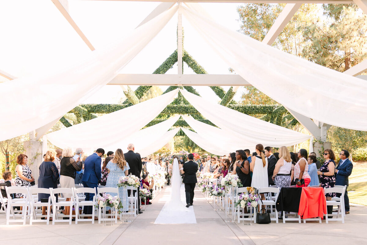 Outdoor wedding ceremony under white drapes and greenery canopy. Guests seated on white chairs, stand as bride and dad walk down the aisle. Elegant, joyful setting.