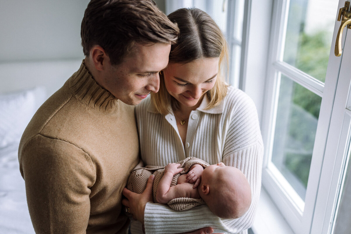 mom in off-white sweater holding newborn wrapped in tan blanket standing next to dad in tan sweater by window