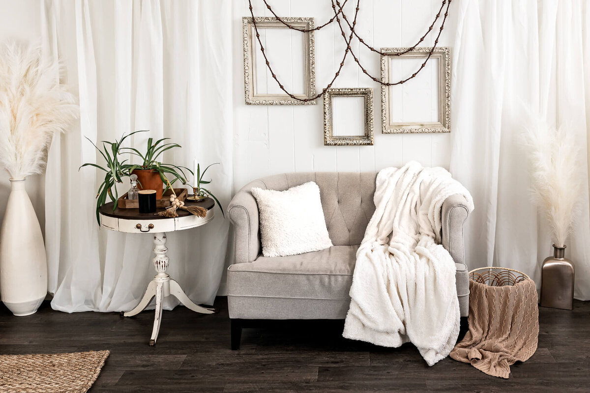 A cozy living room features a gray loveseat with a plush white blanket and pillow. Empty ornate frames hang above. A round side table holds plants and books. Tall vases with pampas grass decorate the space, set against a backdrop of white curtains.