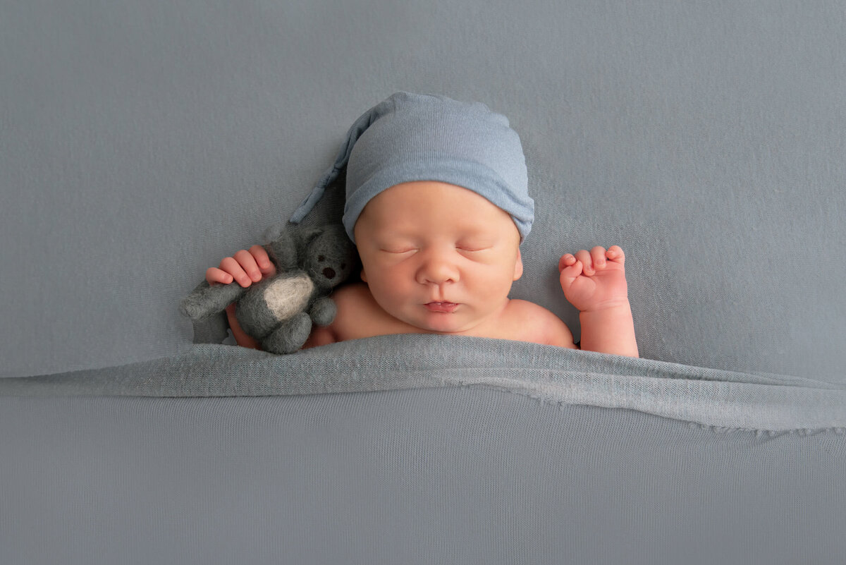 Newborn baby boy wearing a blue sleepy cap on a blue background holding a blue teddy for his Burlington, Ontario newborn photography session.