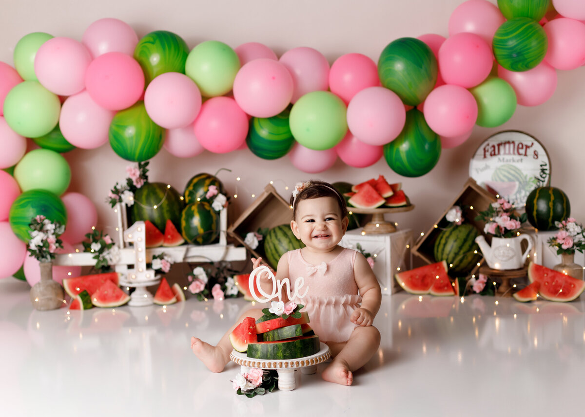 Watermelon themed smash in West Palm Beach and Loxahatchee portrait studio. Baby girl is wearing a soft pink romper sitting in front of sliced watermelon's. In the background is a soft pink and green balloon garland with rustic watermelon accents and wheelbarrow. Baby is looking at the camera smiling.