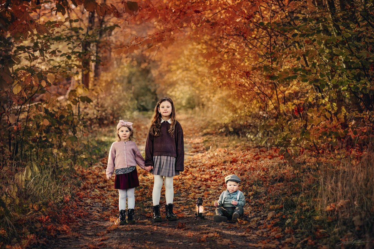 Syskonfotografering utomhus i vackra höstfärger. Två systrar hand  i hand på en grusväg och lillebror sitter på marken bredvid dom.