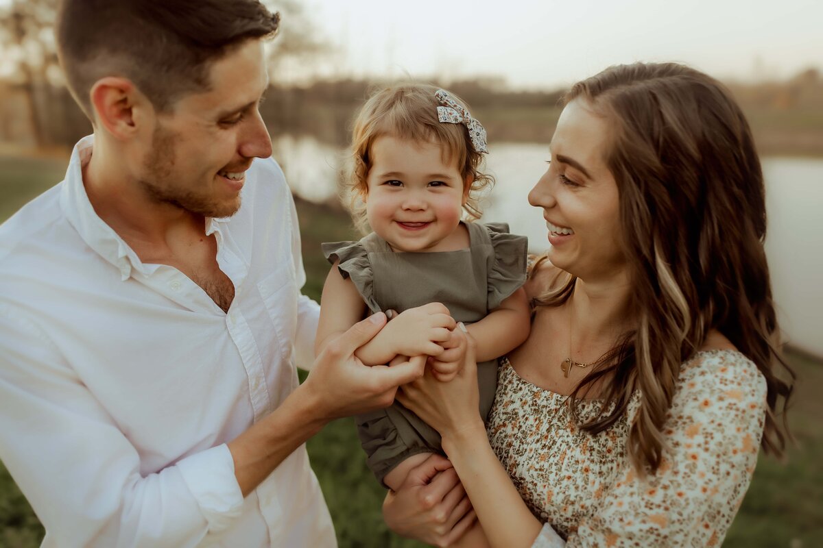 A man and woman smile at a toddler they are holding between them outdoors near a body of water.