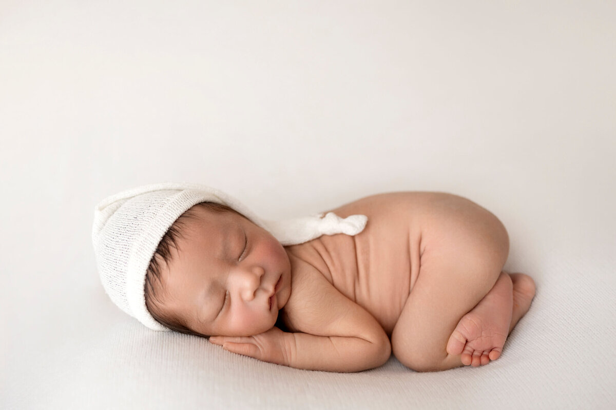 Peacefully sleeping newborn baby curled up on a white blanket, wearing a white knit hat with a small knot on top.