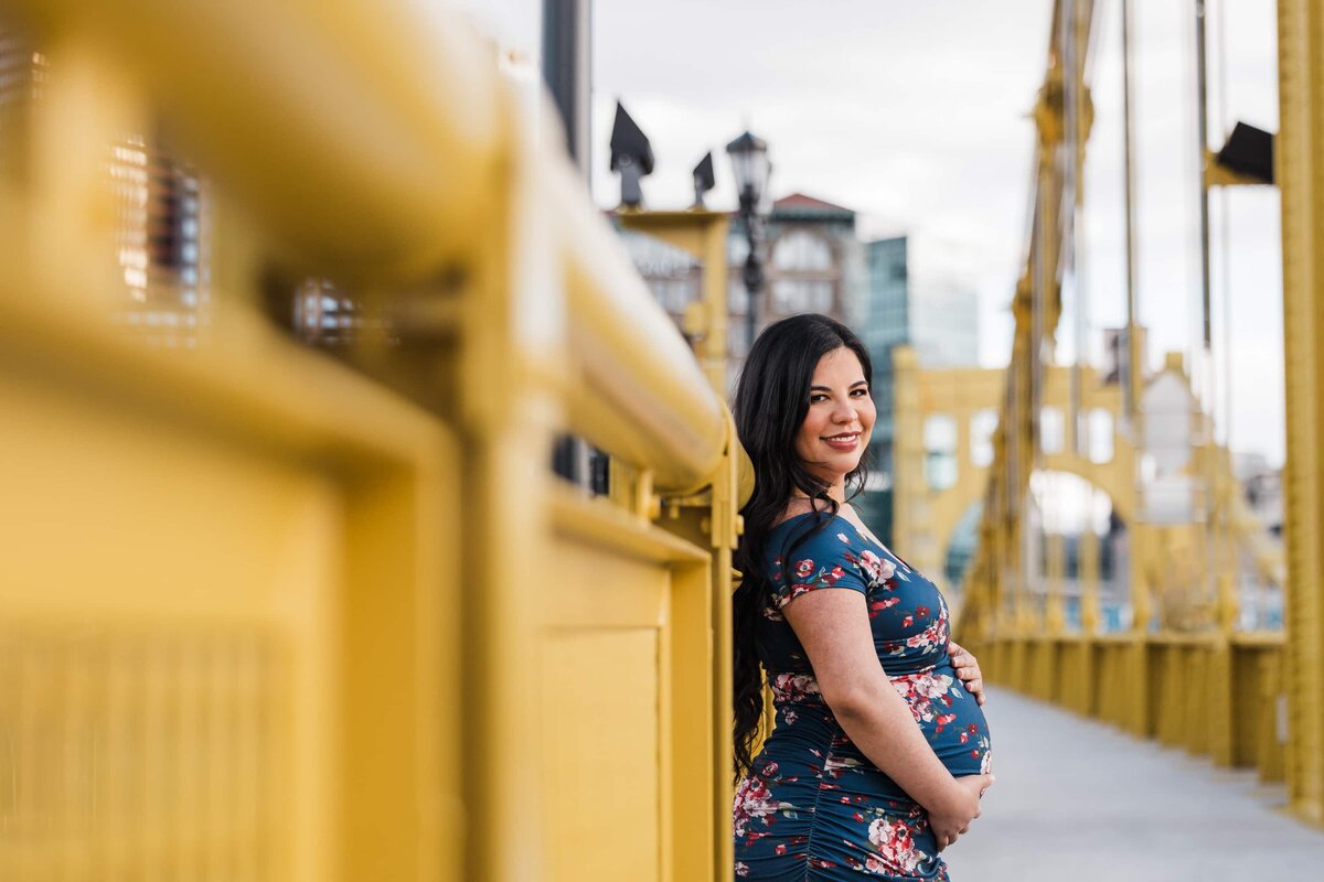 A smiling woman in a floral dress, capturing her maternity moment next to a yellow railing with industrial structures in the Pittsburgh background.