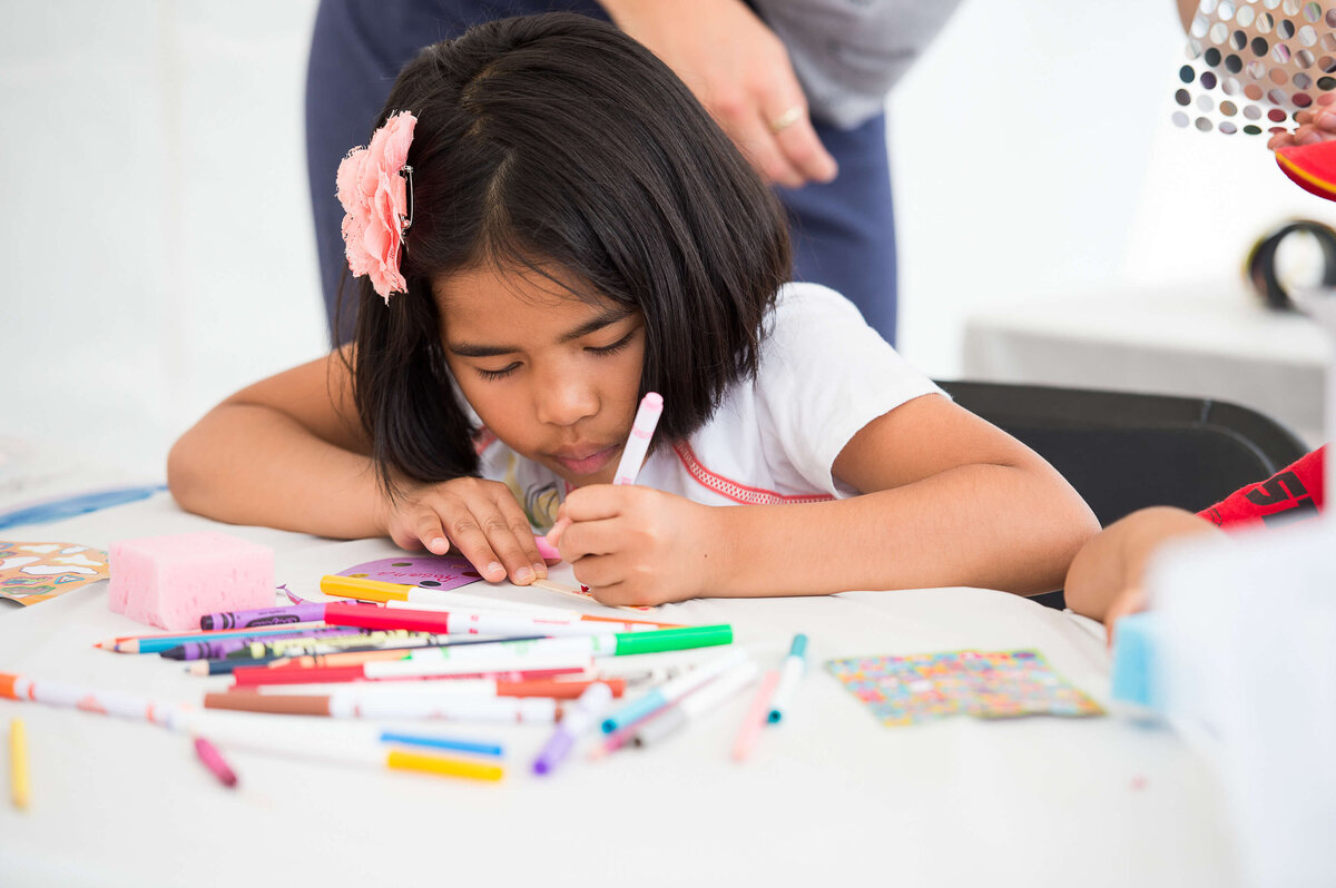 Ottawa event photos showing a little girl with a pink flower in her hair drawing at an activity station as part of a corporate children's event.  Captured by JEMMAN Photography COMMERCIAL