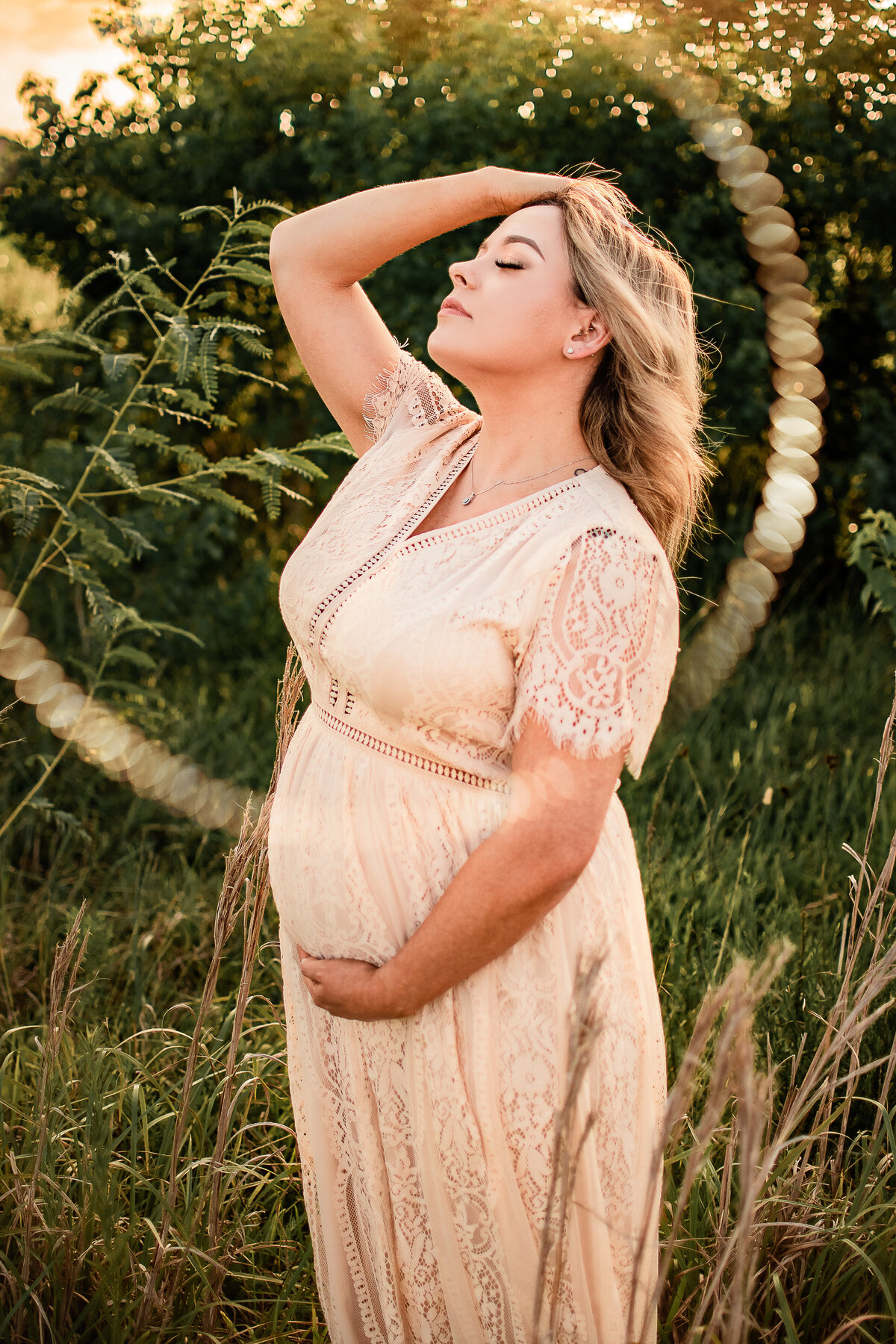 An expectant mother leans her head back and holds her belly with a golden sunset behind her.