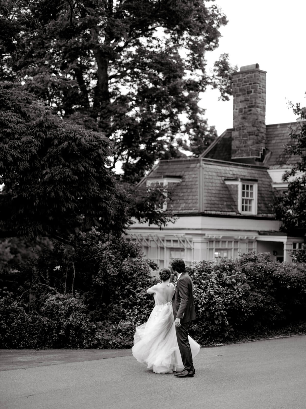 The bride and the groom are standing in front of a big house with a huge tree beside it, while their backs are turned.
