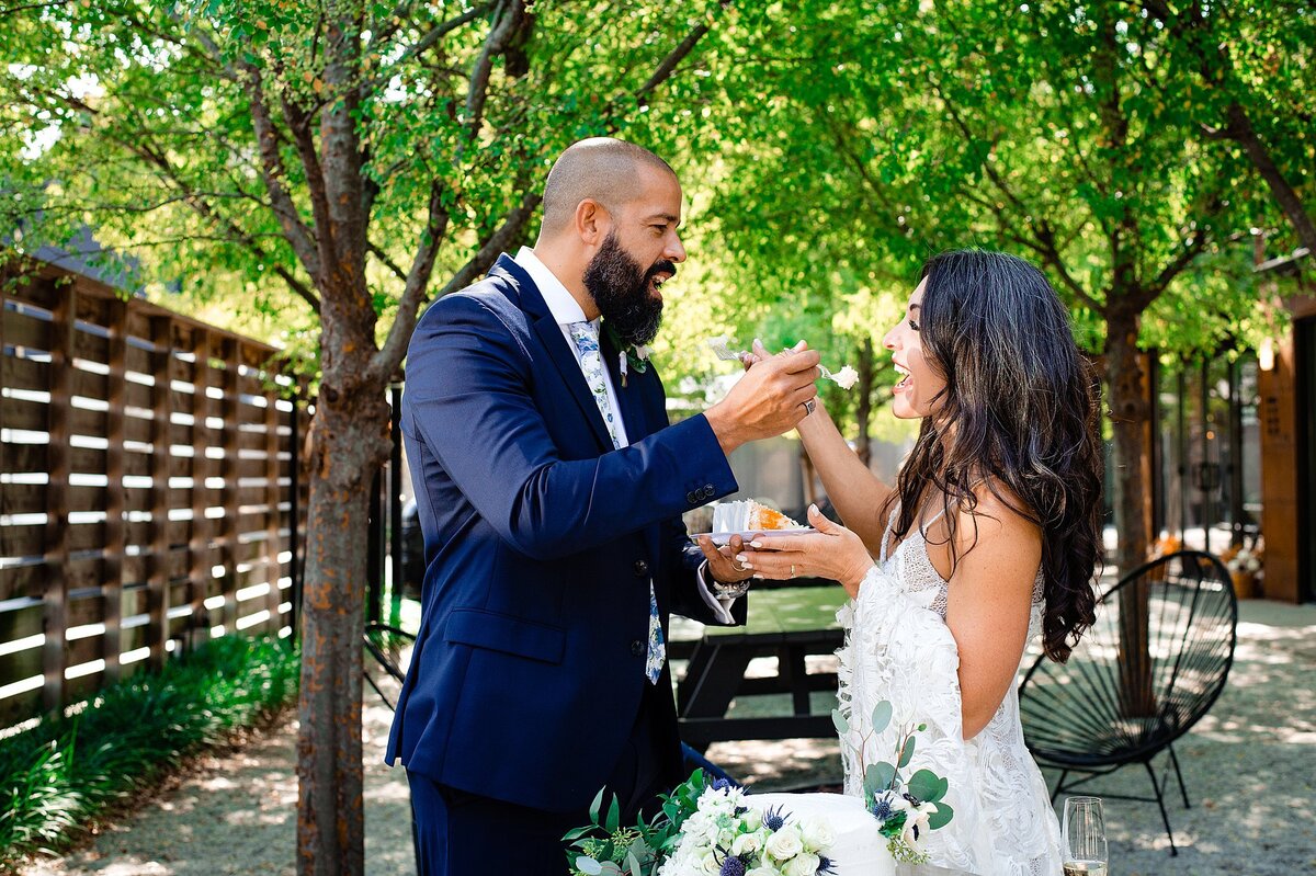 The groom, wearing a dark blue suit, holds up a forkful of cake to the bride who is holding a plate of wedding cake at their elopement at BODE Nashville.
