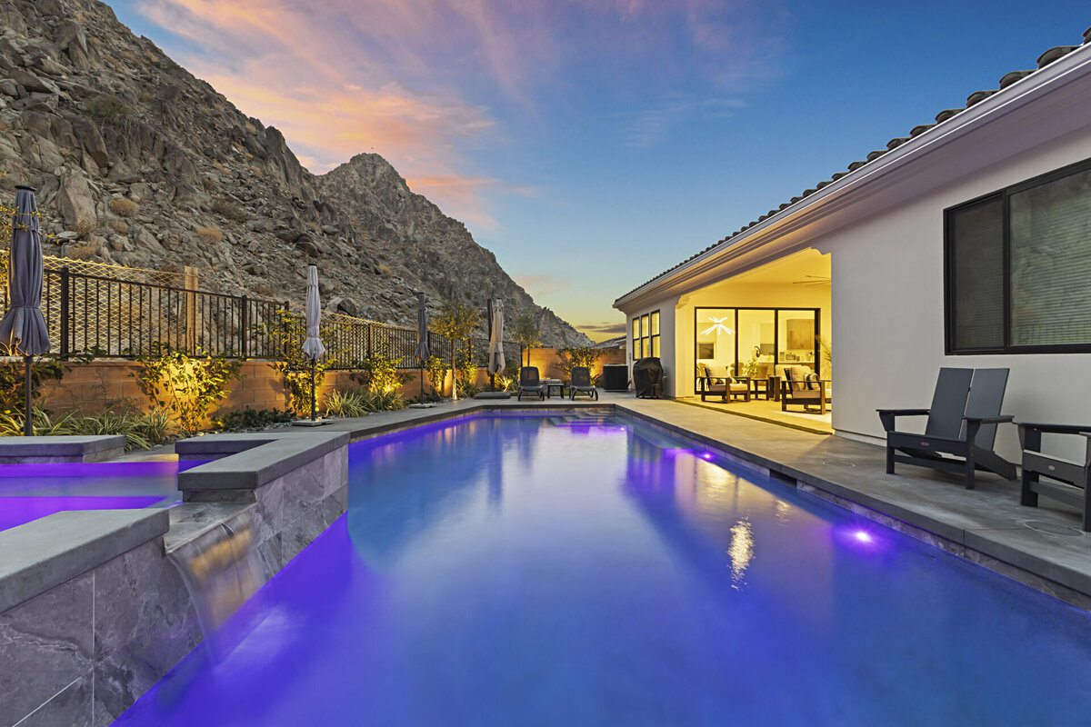 Backyard of desert home with lights on inside and a pool with purple lights at twilight
