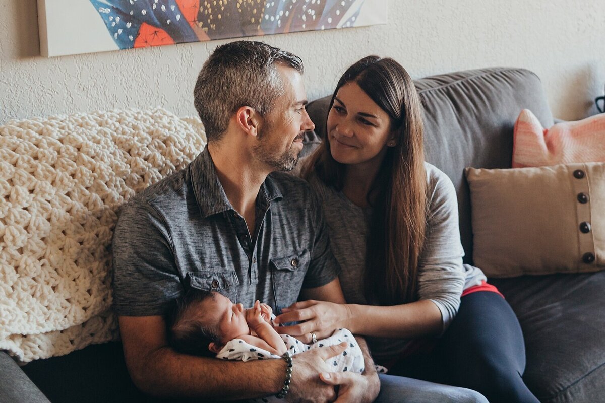 a photo of a mom and dad sitting on a couch looking at each other while dad is holding a newborn baby