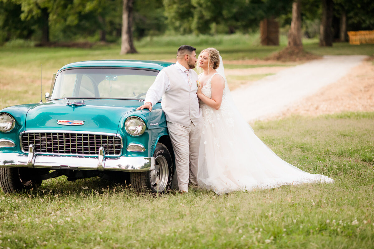 Bride and groom leaning on classic car and leaning toward each other