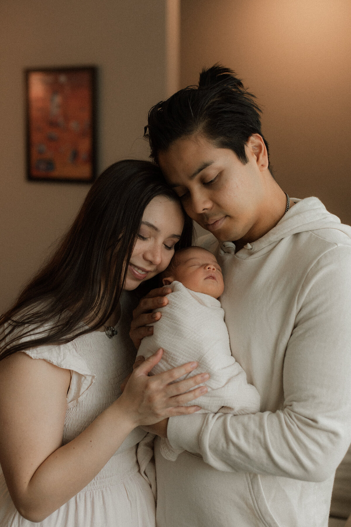 A young couple standing closely together, lovingly gazing at their swaddled newborn cradled in the dad's arms.