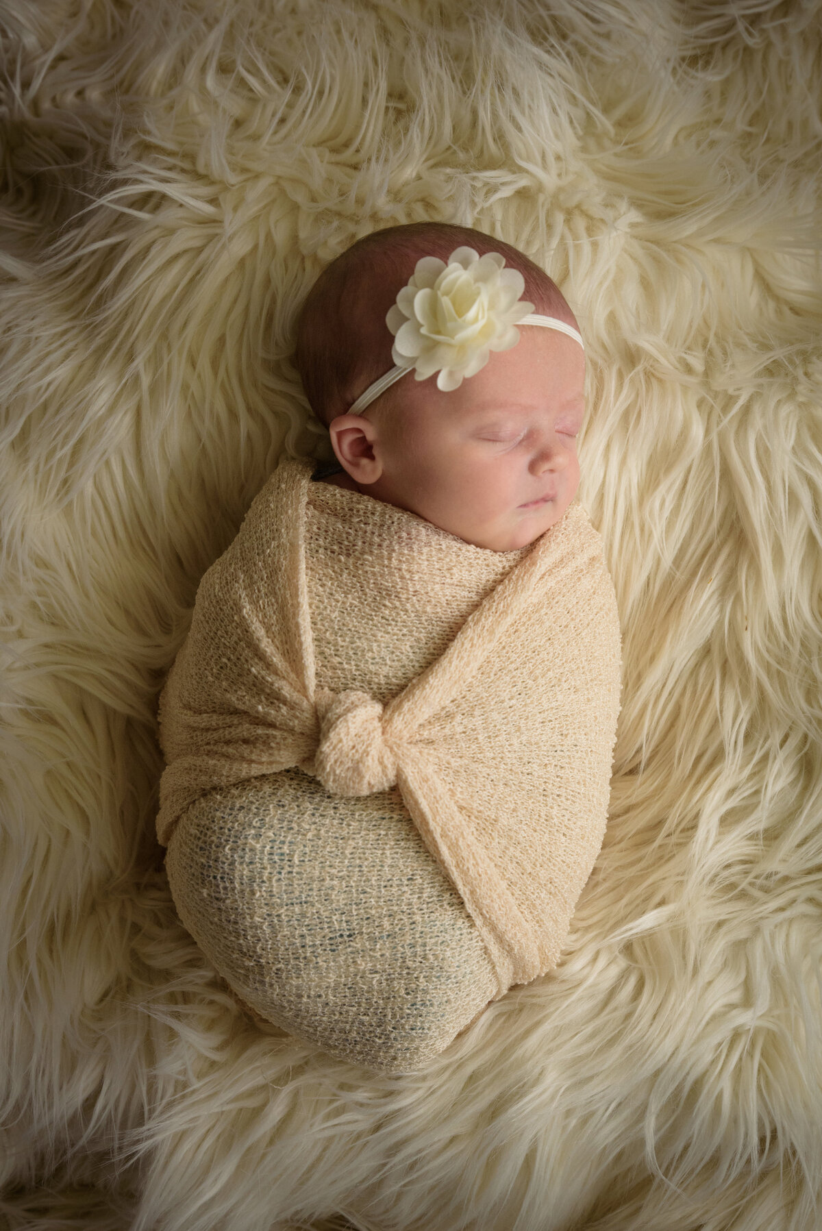 Newborn baby girl in a cream colored wrap and a cream flower sleeping on a fur blanket in her home in Green Bay, Wisconsin