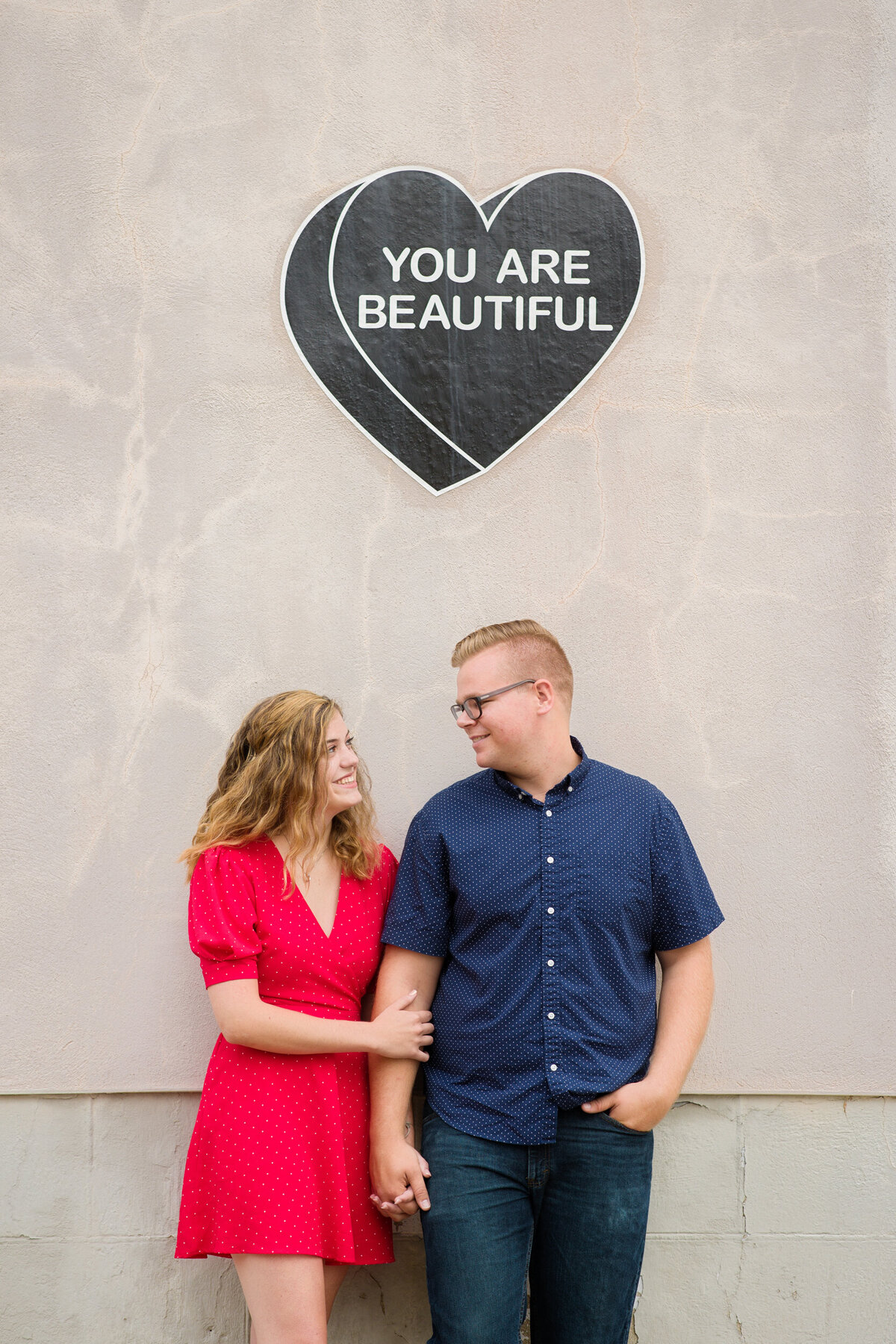 Romantic engagement photo of a couple standing under a 'You Are Beautiful' mural in downtown Manayunk, capturing a vibrant and uplifting moment with artistic charm.