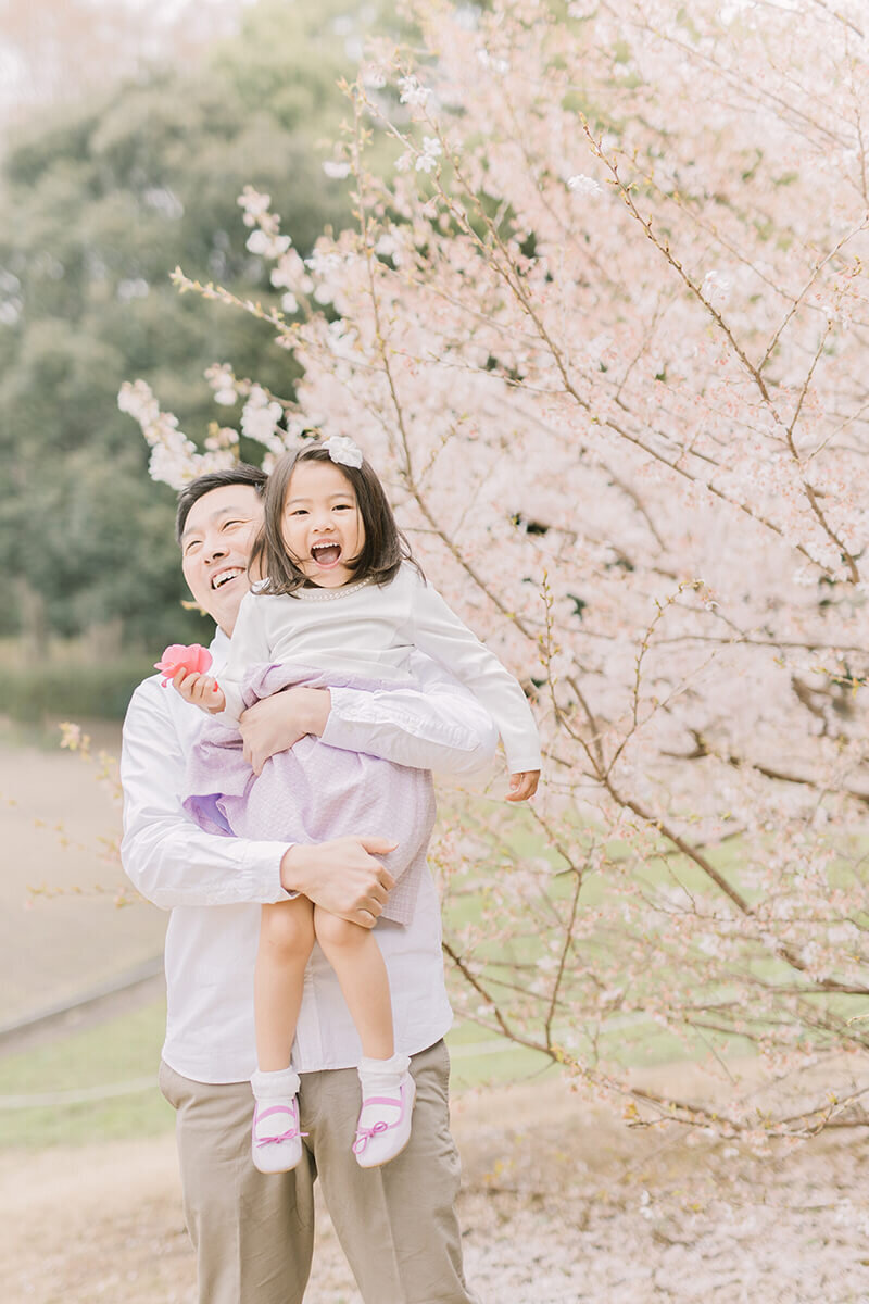 女の子を桜の木の横で抱っこして、二人とも満面の笑みで笑っている可愛らしい家族写真。相模原市の公園で桜の時期に撮影。